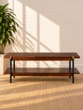 Load image into Gallery viewer, Image 3 of 5: A rectangular wooden coffee table with a dark brown top and lower shelf, supported by black metal legs with brass accents, set against a white brick wall with window shadows and a potted plant.
