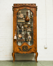 Load image into Gallery viewer, An ornate vintage antique Georgian wooden display cabinet with glass doors, featuring carved details and decorative objects inside, set against a white brick wall.
