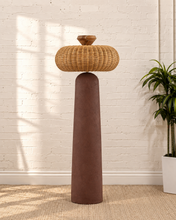 Load image into Gallery viewer, Chandria floor lamp with tall tapered brown fiber cement base, rounded woven rattan shade, and faceted decorative finial.
