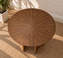Load image into Gallery viewer, A round wooden coffee table with a starburst inlay pattern, shown on a woven jute rug next to a white brick wall and a potted plant.
