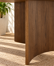 Load image into Gallery viewer, A close-up of a dark brown wooden table base with a curved apron and visible wood grain, set on a woven jute rug in a bright room with a white brick wall and a potted plant in the background.
