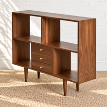Load image into Gallery viewer, Side-angle view of a mid-century modern walnut shelving unit with open cubbies, three center drawers, brass pulls, and tapered legs.
