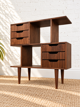 Load image into Gallery viewer, Mid-century modern wooden storage cabinet with six drawers and open shelving, shown on a woven rug against a white brick wall.
