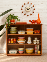 Load image into Gallery viewer, A mid-century modern wooden display shelf filled with retro patterned ceramic dishes, mugs, books, and decorative vases, set against a white brick wall with a starburst wall clock.

