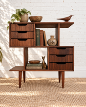 Load image into Gallery viewer, Mid-century modern wooden console table with multiple drawers, open shelving, and tapered legs, styled with books, a plant, a bowl, a vase, and decorative objects against a white brick wall and woven rug.
