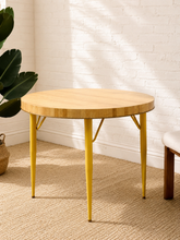 Load image into Gallery viewer, Vintage round Formica dining table with light wood-look top, striped edge detail, and slender mustard yellow metal legs.
