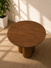 Load image into Gallery viewer, A round wooden side table with a medium brown wood grain finish and three cylindrical legs, styled on a textured beige rug against a white brick wall with soft window shadows.
