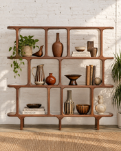 Load image into Gallery viewer, A mid-century modern wooden open shelving unit with a warm brown finish, styled with various ceramic vases, books, and a potted plant against a white brick wall background.

