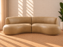 Load image into Gallery viewer, Image 2 of 5: A curved sofa upholstered in tan leather, set against a white brick wall with warm sunlight casting windowpane shadows, on a woven jute rug.
