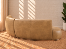 Load image into Gallery viewer, Image 4 of 5: A curved tan leather sofa shown from the back in a styled room with a white brick wall, sunlight shadows, a woven rug, and a potted plant.
