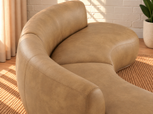 Load image into Gallery viewer, Image 5 of 5: A curved, modern accent chair upholstered in tan leather, shown in a brightly lit room with a white brick wall, a woven rug, and a potted plant.
