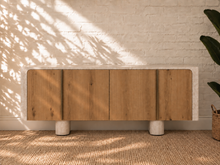 Load image into Gallery viewer, Marlee four-door credenza with natural oak veneer doors, faux travertine surround, interior shelving, and two rounded pedestal legs.
