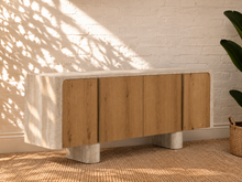 Load image into Gallery viewer, Marlee four-door credenza with natural oak veneer doors, faux travertine surround, interior shelving, and two rounded pedestal legs.
