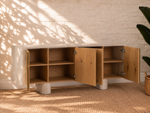 Load image into Gallery viewer, Marlee four-door credenza with natural oak veneer doors, faux travertine surround, interior shelving, and two rounded pedestal legs.
