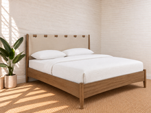 Load image into Gallery viewer, A modern wooden bed frame with a beige upholstered headboard, styled with white bedding and pillows in a bright, sunlit bedroom with a potted plant.
