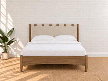 Load image into Gallery viewer, A modern wooden bed frame with a beige upholstered headboard featuring leather tie details, styled with white bedding and set against a white brick wall with a potted plant on the left.
