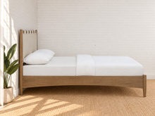 Load image into Gallery viewer, A modern single bed with a light oak wood frame and a beige canvas headboard, dressed in white bedding, set against a white brick wall with a potted plant on a woven rug.
