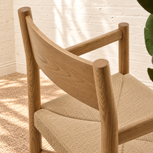 Load image into Gallery viewer, Amelie natural solid oak dining armchair with woven paper-rope seat, curved wood backrest, slim armrests, and X-shaped stretcher
