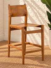 Load image into Gallery viewer, Nova counter stool in natural brown genuine leather with a tall teak wood frame, leather seat and backrest, footrest, and buckle strap detail.
