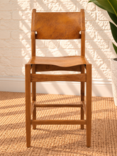 Load image into Gallery viewer, Nova counter stool in natural brown genuine leather with a tall teak wood frame, leather seat and backrest, footrest, and buckle strap detail.
