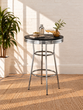 Load image into Gallery viewer, Image 2 of 5: A round bar-height table with a black top and chrome metal legs, styled with a bottle, glasses, and a small plant against a white brick wall.
