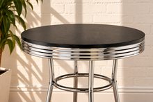 Load image into Gallery viewer, Image 3 of 5: A round bar table with a black faux-wood top and a chrome metal swivel base, set against a white brick wall with soft shadows.
