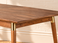 Load image into Gallery viewer, Image 3 of 4: A mid-century modern wooden dining table with tapered legs and brass metal accents, set in a room with a white brick wall, a potted plant, and a woven rug.
