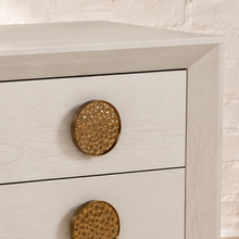 Load image into Gallery viewer, Phoebe three-drawer nightstand in cream white ash veneer with six round textured golden metal handles and recessed plinth base.

