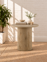 Load image into Gallery viewer, Image 2 of 4: A round side table with a fluted pedestal base in a light natural wood finish, styled with books, a bowl, a candle, and a small vase of greenery against a white brick wall.
