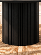 Load image into Gallery viewer, Image 4 of 4: A round black dining table with a fluted pedestal base, set on a woven beige rug against a white brick wall with warm sunlight and shadow patterns.
