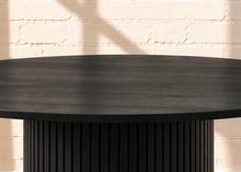 Load image into Gallery viewer, Image 3 of 4: A round black dining table with a fluted pedestal base, set on a woven beige rug against a white brick wall with warm sunlight and shadow patterns.
