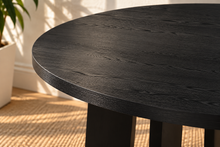 Load image into Gallery viewer, Image 4 of 4: A close-up of a round dining table with a dark charcoal wood grain finish and a thick overhang lip, shown in a brightly lit room with a potted plant and woven rug in the background.
