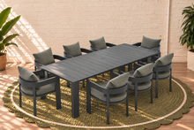 Load image into Gallery viewer, Image 1 of 4: An 9-piece outdoor dining set featuring a rectangular slatted table and matching armchairs with dark gray frames and light gray cushions, arranged on a round woven jute rug in front of a white brick wall.
