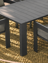 Load image into Gallery viewer, Image 3 of 4: Dark gray outdoor dining table with matching armchairs on a textured beige woven rug, shown in a real patio setting with natural sunlight and shadows.
