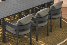 Load image into Gallery viewer, Image 4 of 4: An 9-piece outdoor dining set featuring a rectangular slatted table and matching armchairs with dark gray frames and light gray cushions, arranged on a round woven jute rug in front of a white brick wall.
