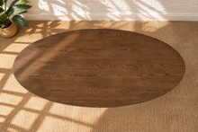 Load image into Gallery viewer, An oval wooden dining table with a rich medium-brown finish and visible wood grain, shown on a textured beige rug with soft window shadow patterns.
