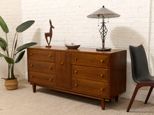 Load image into Gallery viewer, A mid-century modern wooden sideboard or credenza with six drawers, styled in a room setting with a white brick wall, a potted plant, a black chair, and a table lamp on top.
