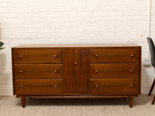Load image into Gallery viewer, Mid-century modern wooden sideboard with six drawers and a central door, featuring brass knobs, set against a white brick wall.
