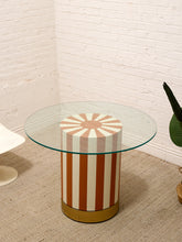 Load image into Gallery viewer, A modern round side table with a clear glass top and a cylindrical pedestal base featuring vertical rust-orange and cream stripes with a metallic gold circular base, set in a room corner with a white brick wall and beige carpet.
