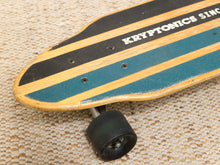 Load image into Gallery viewer, Close-up of a longboard skateboard resting on a woven beige mat, showing the deck with alternating black and natural wood stripes, teal grip tape, white KRYPTOMICS branding, and a black wheel.
