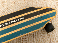 Load image into Gallery viewer, A close-up of a wooden longboard skateboard with teal and black striped grip tape, showing the text 'SKATEBOARDS SINCE 1965' and a black wheel, resting on a textured beige woven mat.
