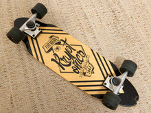 Load image into Gallery viewer, A longboard skateboard with a natural wood deck, black graphic stripes, and a 'Puppet since 1983' logo, resting on a beige woven rug.

