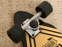 Load image into Gallery viewer, Close-up of a skateboard deck and trucks, showing black wheels, a silver metal truck, and a light wood deck with black graphic stripes.
