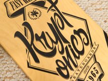 Load image into Gallery viewer, Close-up of a light natural wood skateboard deck with black engraved branding reading 'Puppet enics' and 'Skateboards' on a woven beige surface.
