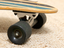Load image into Gallery viewer, Close-up of a skateboard's rear wheel and truck assembly resting on a textured beige woven surface. The skateboard has a worn light wood deck with teal grip tape and black stripes, black wheels, and a white truck.
