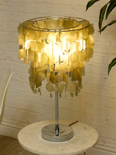 Load image into Gallery viewer, A table lamp with a circular shade made of overlapping translucent gold discs, illuminated from within, mounted on a chrome stand on a round stone side table against a white brick wall.
