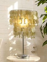 Load image into Gallery viewer, A table lamp with a circular shade made of layered translucent pale yellow shell-like pieces, mounted on a slim silver metal stand with a round base, placed on a light stone side table against a white brick wall with green leaves at the side.
