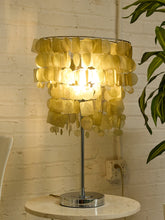 Load image into Gallery viewer, A table lamp with a circular shade made of overlapping translucent yellow-gold discs, glowing from within on a silver metal stand, placed on a round white table against a white brick wall.
