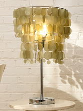 Load image into Gallery viewer, A table lamp with a circular shade made of overlapping translucent gold-toned discs, mounted on a slim chrome stand with a round base, set on a round stone table against a white brick wall.
