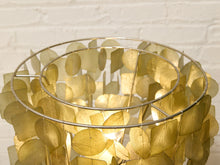 Load image into Gallery viewer, A close-up of a decorative lampshade made of overlapping translucent gold-toned leaf discs mounted on a thin circular metal frame, glowing from within against a white brick wall background.
